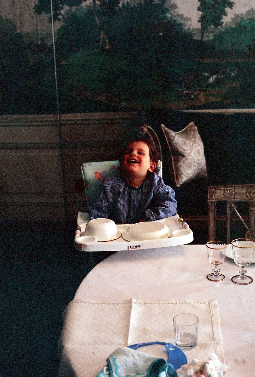 Carlo Acutis sorrindo à mesa posta, com um quadro de paisagem na parede atrás dele